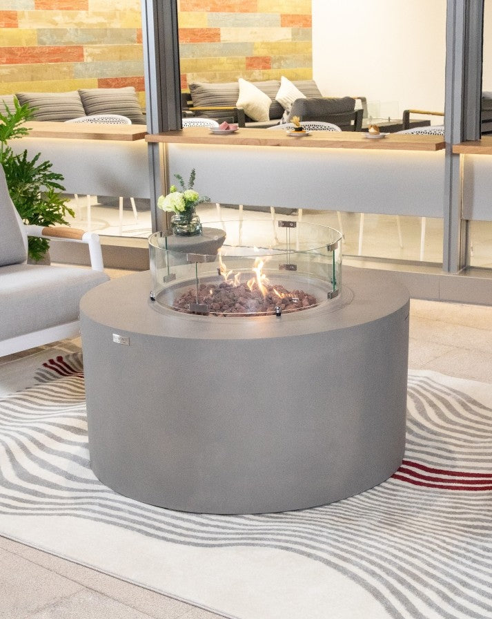 Elementi Pacaya Round Hidden Propane Tank Fire Pit Table in light grey GFRC concrete, featuring a warm, flickering flame over natural lava rocks, elegantly enclosed by a glass wind guard for a modern and inviting outdoor ambiance.