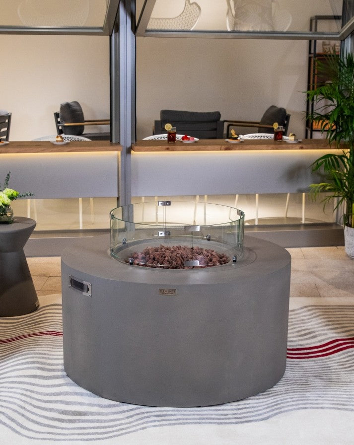 Elementi Pacaya Round Hidden Propane Tank Fire Pit Table in light grey GFRC concrete, featuring a stylish glass wind guard and natural lava rocks, set in a modern outdoor lounge for a sophisticated and cozy ambiance.