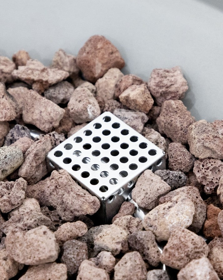 "Close-up of the Elementi Pacaya Round Hidden Propane Tank Fire Pit Table's stainless steel burner nestled among natural lava rocks. This high-quality GFRC concrete fire feature offers a modern aesthetic with durable, heat-resistant materials for a stylish and long-lasting outdoor ambiance.