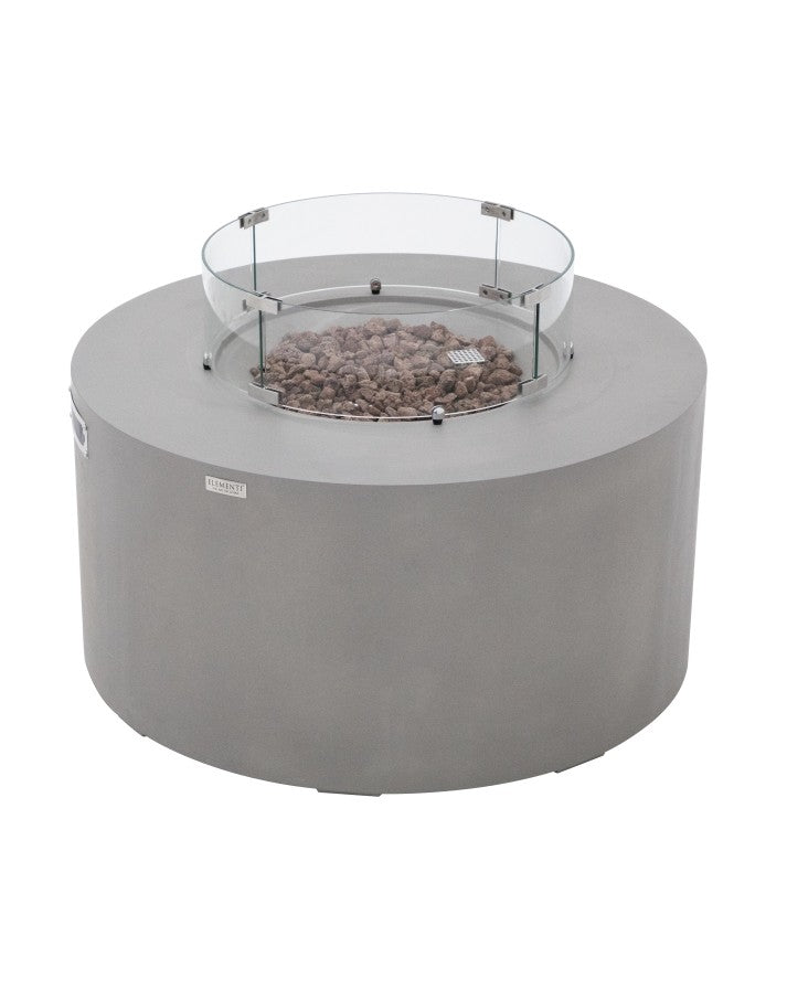 Elementi Pacaya Round Hidden Propane Tank Fire Pit Table in light grey GFRC concrete, showcasing rustic lava rocks and a protective glass wind guard. This elegantly designed fire feature blends durability with modern aesthetics, making it a perfect centerpiece for outdoor ambiance and warmth.