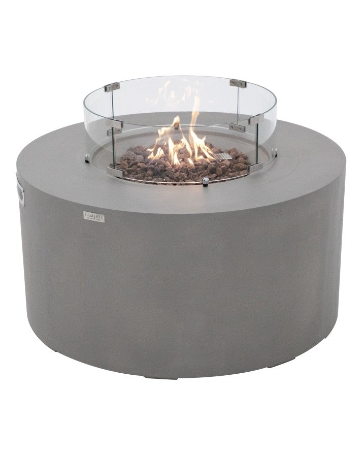 Elementi Pacaya Round Hidden Propane Tank Fire Pit Table in light grey GFRC concrete, glowing with vibrant flames atop rustic lava rocks. The sleek glass wind guard enhances safety while complementing the modern design, making this fire pit a sophisticated centerpiece for any outdoor living space.