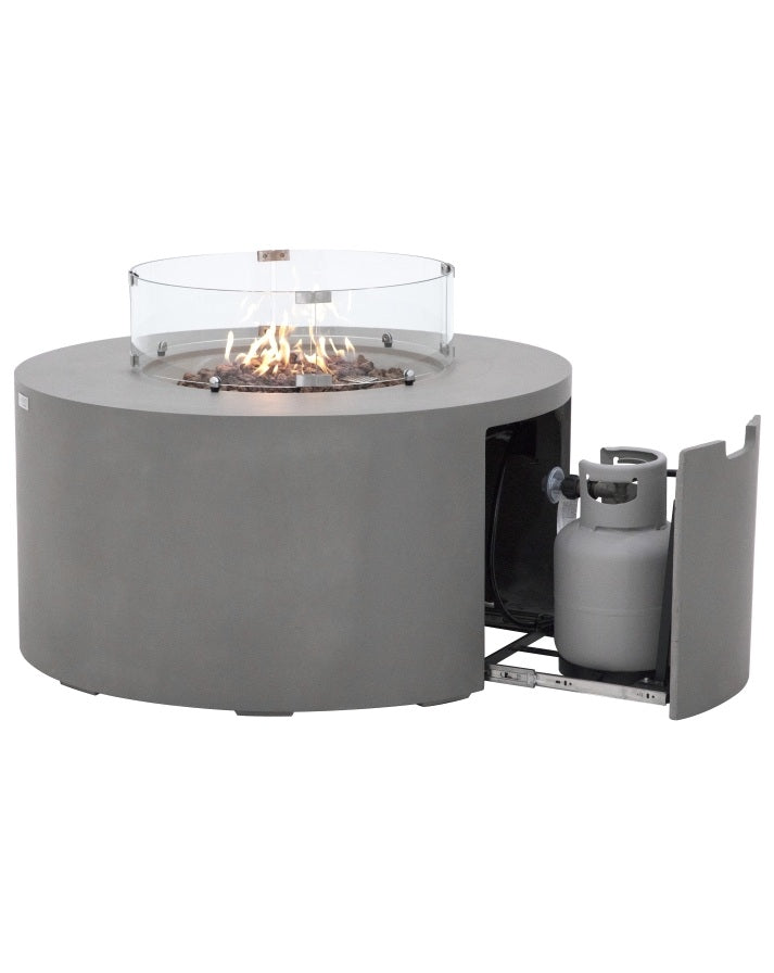 Elementi Pacaya Round Hidden Propane Tank Fire Pit Table in light grey GFRC concrete, featuring a mesmerizing flame over rustic lava rocks. The sleek glass wind guard enhances safety and ambiance, while the discreet pull-out drawer reveals a hidden propane tank compartment for a clean, modern aesthetic.