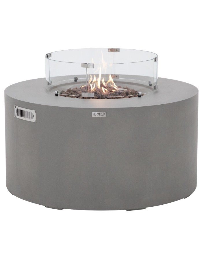 Elementi Pacaya Round Hidden Propane Tank Fire Pit Table in light grey GFRC concrete, elegantly ignited with flames dancing over natural lava rocks. Featuring a stylish glass wind guard, this modern fire table creates a warm and inviting outdoor ambiance while offering a sleek, contemporary design with hidden propane tank storage.