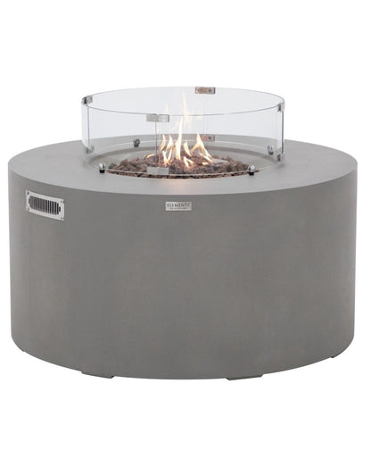 Elementi Pacaya Round Hidden Propane Tank Fire Pit Table in light grey GFRC concrete, elegantly ignited with flames dancing over natural lava rocks. Featuring a stylish glass wind guard, this modern fire table creates a warm and inviting outdoor ambiance while offering a sleek, contemporary design with hidden propane tank storage.