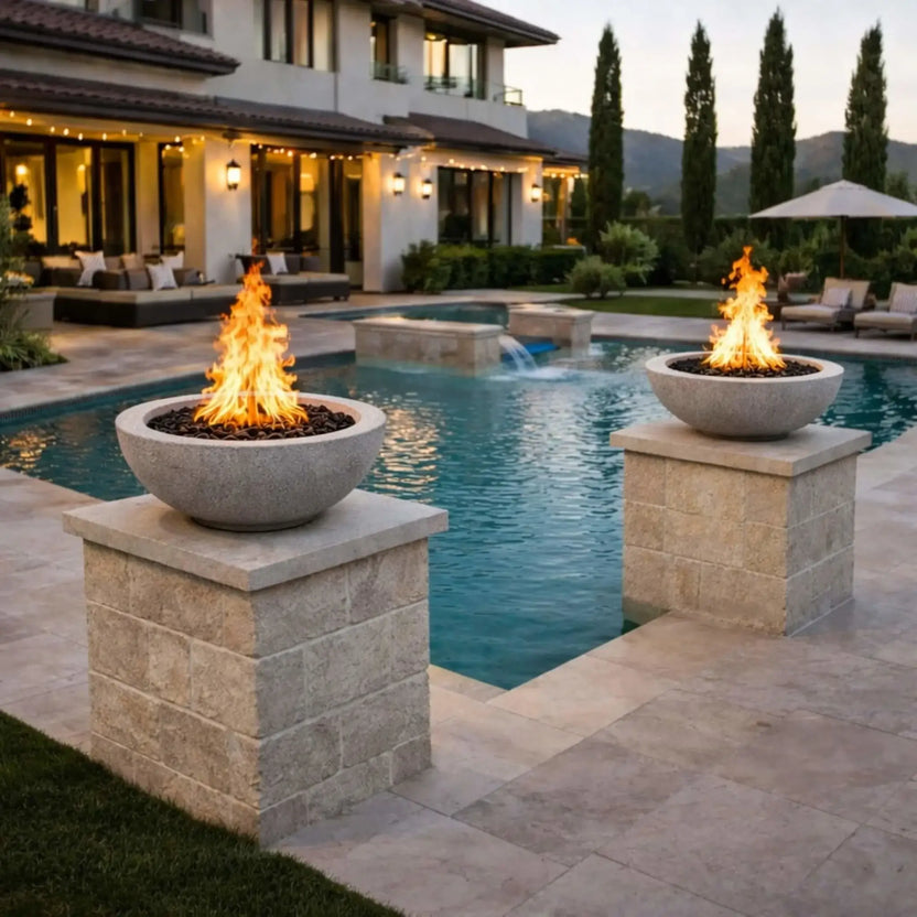 Two Flame Havens round concrete fire bowls in a gray finish set atop stone pedestals beside a luxury swimming pool, with tall vibrant flames reflecting off the water and complementing a modern Mediterranean-style backyard design.