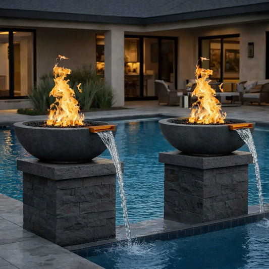 Pair of round black concrete fire and water bowls installed on stone pedestals beside a modern swimming pool, featuring tall gas flames rising from black fire media while water spills smoothly from bronze scuppers into the pool, creating a dramatic fire and water focal point in a contemporary backyard setting.