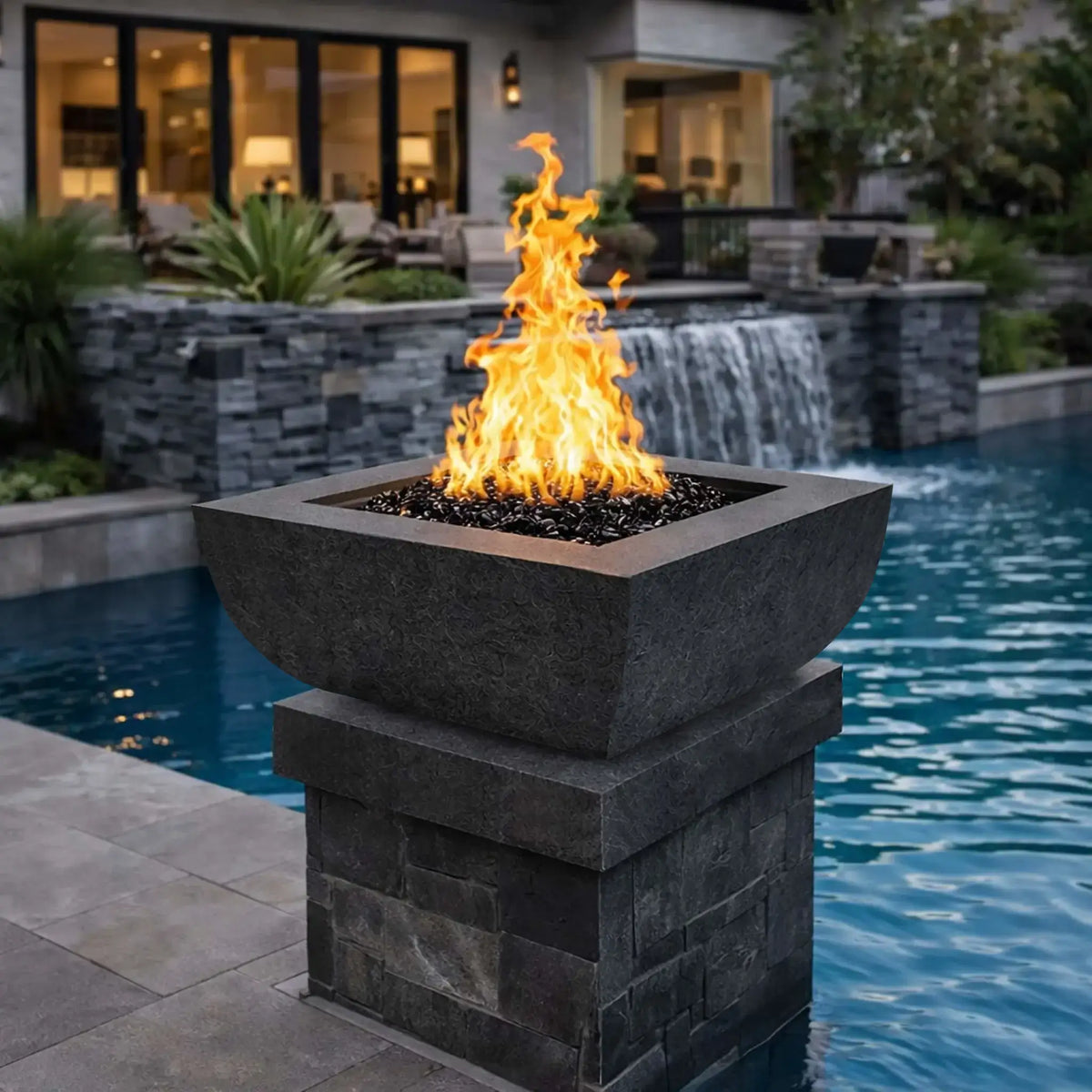 Dark graphite square curved fire bowl from the Flame Havens Serenity Series installed beside a luxury swimming pool. The modern fire bowl sits on a stacked stone pedestal and produces tall yellow flames above black fire glass. In the background, a contemporary home patio, large glass doors, landscaping, and a cascading pool waterfall create an upscale outdoor living setting.