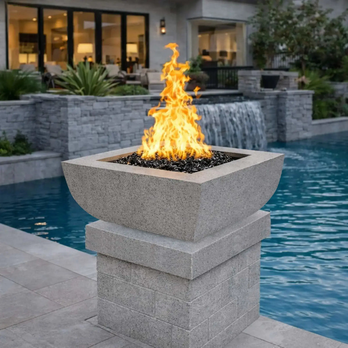 Light gray Serenity Series square curved fire bowl mounted on a stone pedestal next to a modern swimming pool. Bright gas flames rise above black fire glass while a stone pool wall and waterfall spillway flow into the water behind it. The scene includes landscaped plants and a contemporary patio with large windows and outdoor seating.