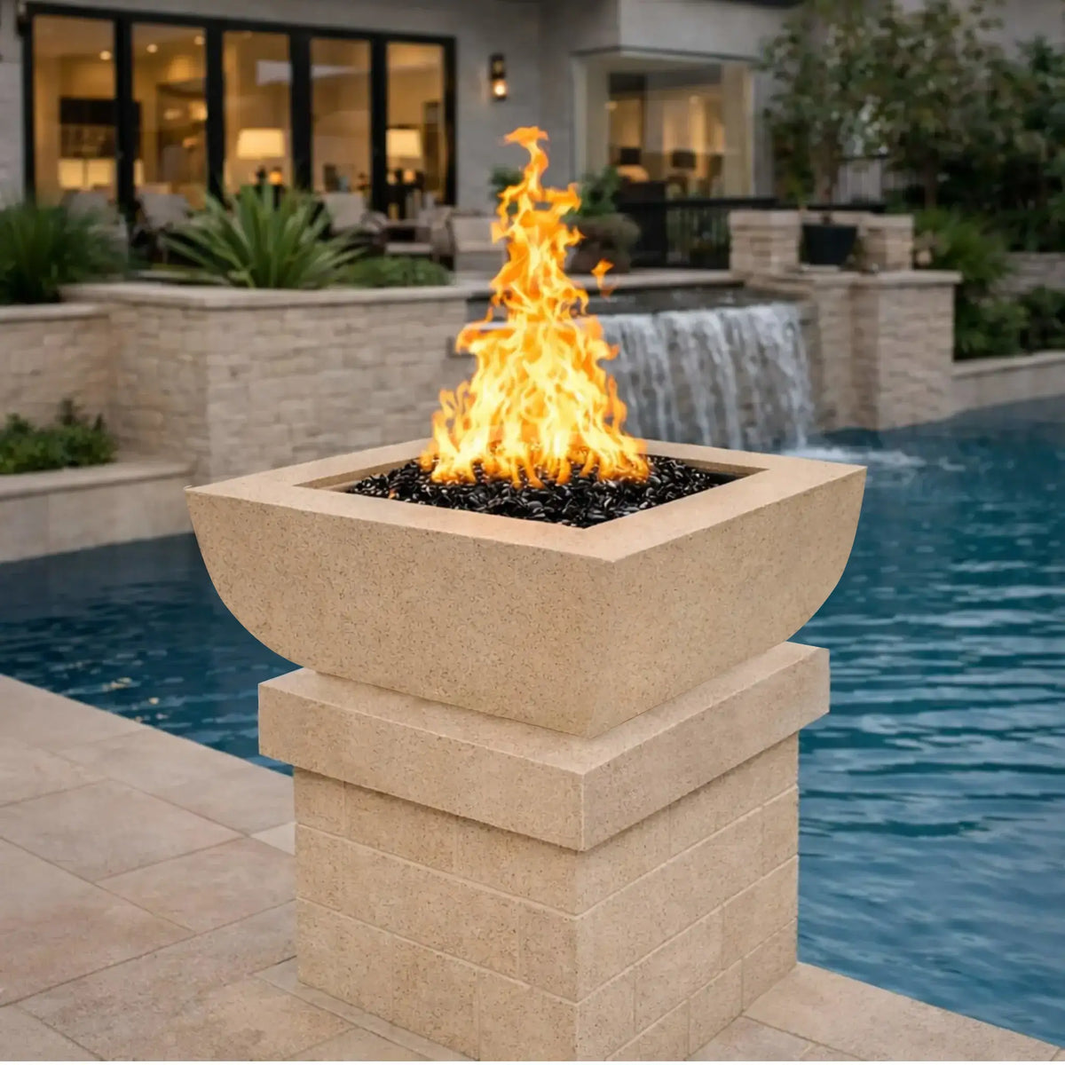 Soft white square curved fire bowl from the Flame Havens Serenity Series displayed in a luxury backyard pool setting. The gas fire bowl burns vibrant flames above dark fire media while positioned on a matching pedestal beside a blue swimming pool. A natural stone wall with a waterfall feature and modern patio lounge area appear in the background.