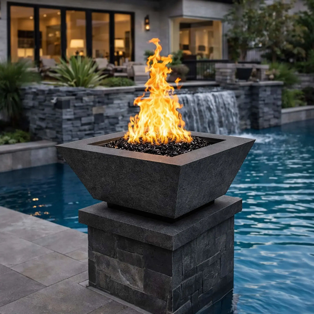 Flame Havens 24 inch black square angular fire bowl mounted on a stone column at the edge of a luxury pool, complemented by a textured stone waterfall backdrop, showcasing reflective black fire glass and a dramatic flame ideal for modern custom pool environments.
