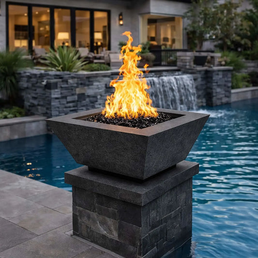 Flame Havens 24 inch black square angular fire bowl mounted on a stone column at the edge of a luxury pool, complemented by a textured stone waterfall backdrop, showcasing reflective black fire glass and a dramatic flame ideal for modern custom pool environments.