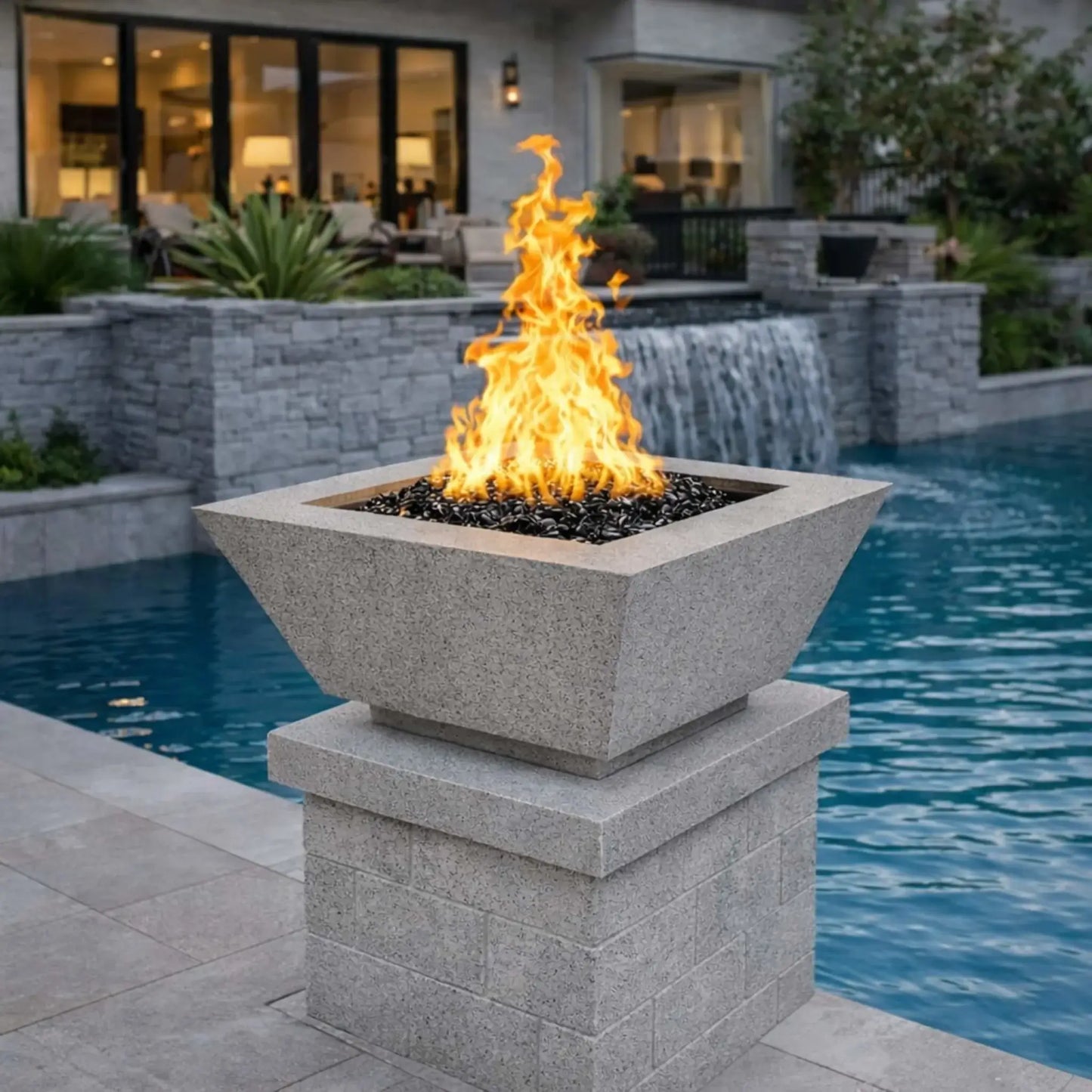 Flame Havens 24 inch square angular fire bowl in gray, set on a stone pillar beside a custom swimming pool with a cascading waterfall, featuring reflective black fire glass and a tall, dynamic flame that adds a refined modern accent to a luxury backyard pool and spa design.