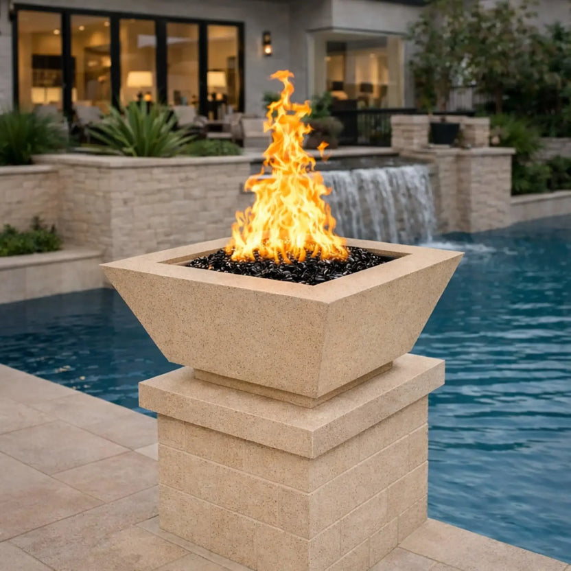 Flame Havens 24 inch white square angular fire bowl displayed on a masonry pedestal along a resort-style pool, framed by a natural stone waterfall and upscale landscaping, with reflective black fire glass creating a bold flame centerpiece for elegant outdoor living spaces.