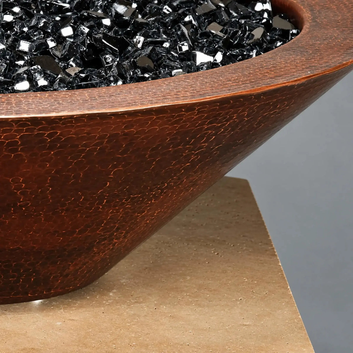 Close-up of the Flame Havens Bella fire and water bowl, showing the dimpled hammered copper finish, wide rounded rim, and glossy black fire glass. Light catches the angular glass pieces and warm copper surface, while the tapered side of the bowl rests on a pale stone pedestal against a gray background.