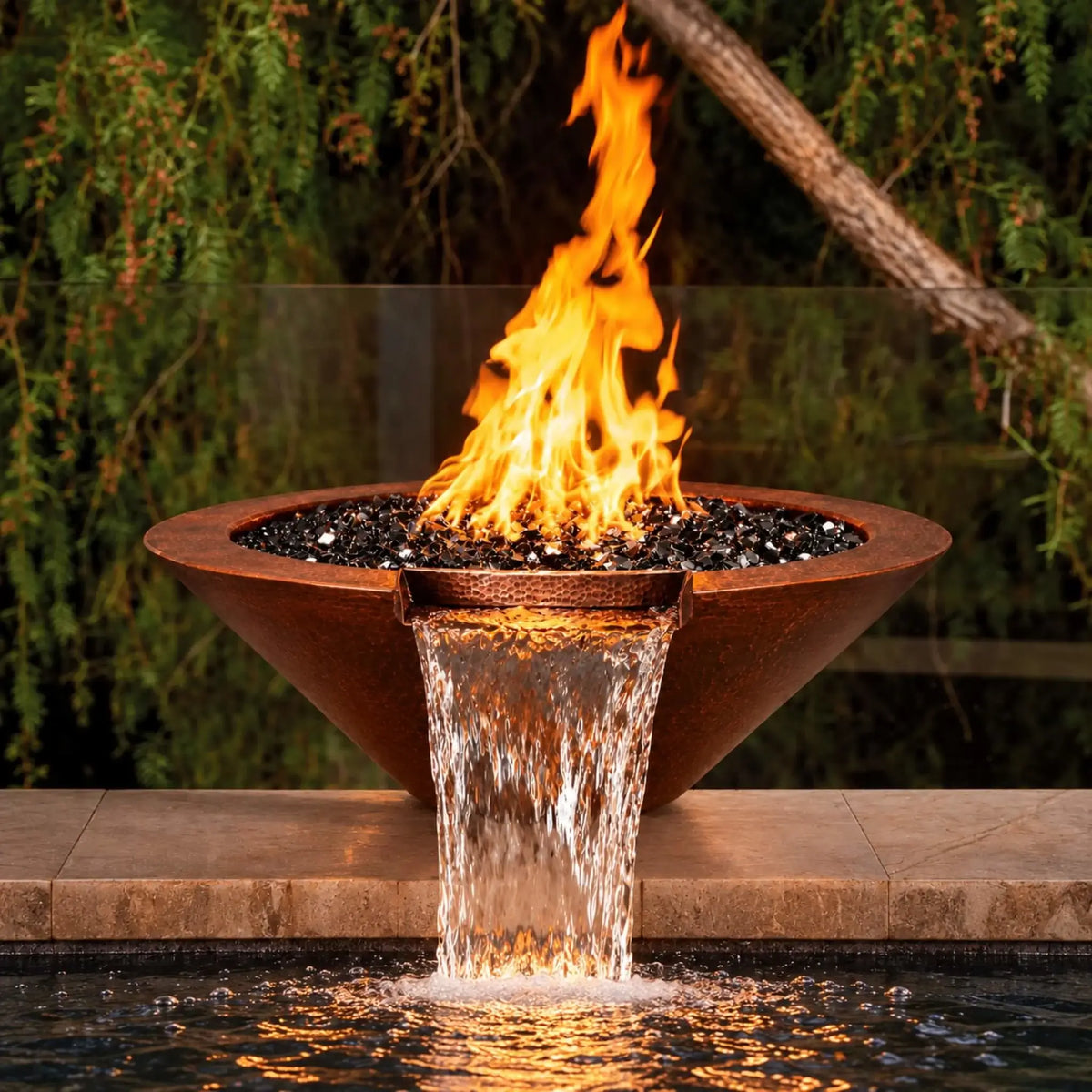 Flame Havens Bella hammered copper fire and water bowl on a tan stone pool ledge, viewed from the front. Tall orange flames rise above reflective black fire glass as a broad sheet of water pours from the copper scupper into the pool. Firelight glows across the waterfall and rippling water, with leafy trees behind the bowl.