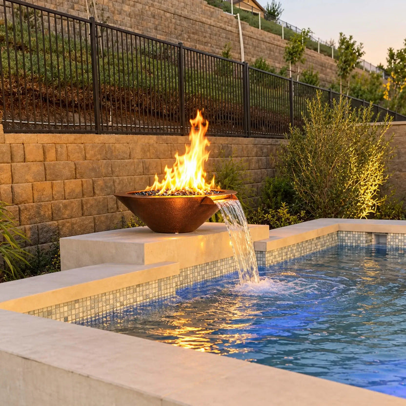 Flame Havens Bella fire and water bowl in hammered copper sits on a raised cream-colored pedestal beside a blue mosaic-tiled pool. Bright flames rise from black fire glass, and water arcs from the copper spillway into the pool. Shrubs, tiered stone retaining walls, and a black metal fence frame the backyard setting in warm evening light.