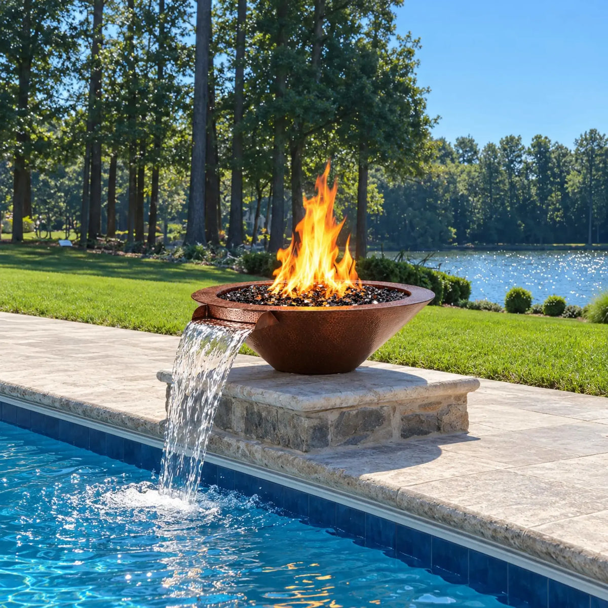 Flame Havens Bella hammered copper fire and water bowl on a low stone pedestal beside a bright blue swimming pool. Orange flames rise from black fire glass while a wide waterfall flows from the front copper scupper. Pale stone paving, a green lawn, mature trees, and a sunlit lake surround the poolside installation.