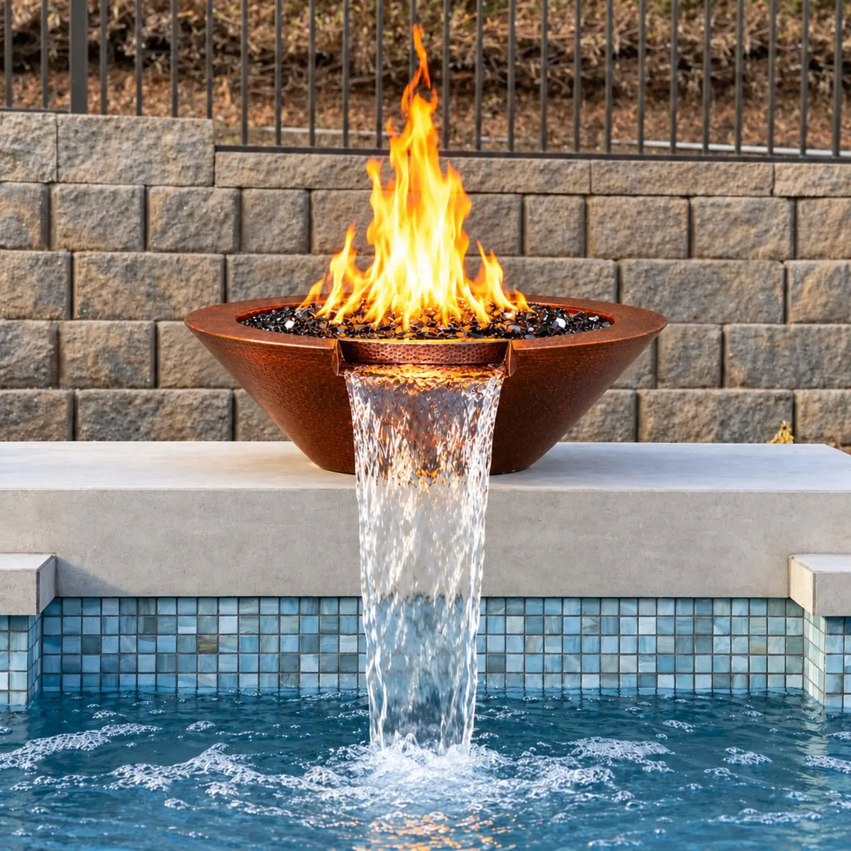 Front view of the Flame Havens Bella hammered copper fire and water bowl on a pale pool ledge. Tall yellow-orange flames burn above glossy black fire glass, and the centered copper scupper releases a broad waterfall into the blue pool below. Blue mosaic waterline tiles, a stone block wall, and black metal fencing complete the setting.
