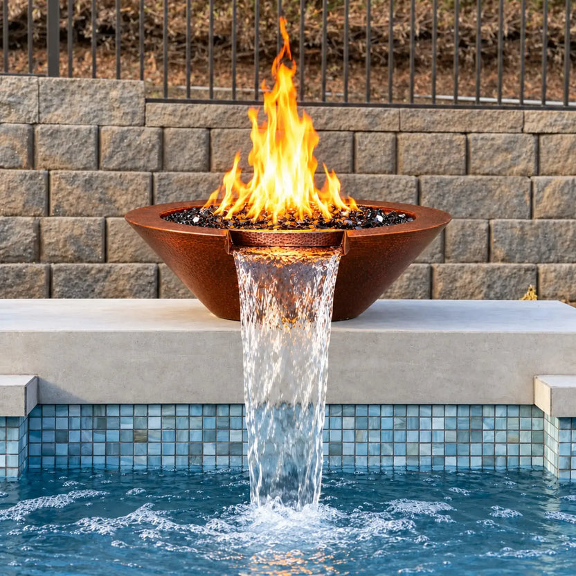Front view of the Flame Havens Bella hammered copper fire and water bowl on a pale pool ledge. Tall yellow-orange flames burn above glossy black fire glass, and the centered copper scupper releases a broad waterfall into the blue pool below. Blue mosaic waterline tiles, a stone block wall, and black metal fencing complete the setting.