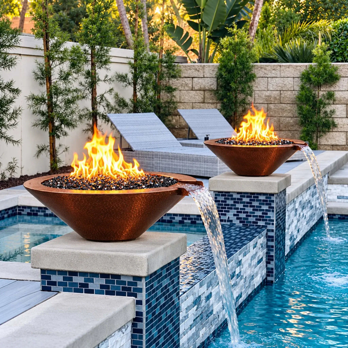 Two Flame Havens Bella fire and water bowls in hammered copper line a pool on pillars faced with blue mosaic tile and topped with pale stone caps. Orange flames dance above reflective black fire glass as water pours from each copper scupper into the pool. Gray woven lounge chairs and tropical greenery fill the patio behind them.