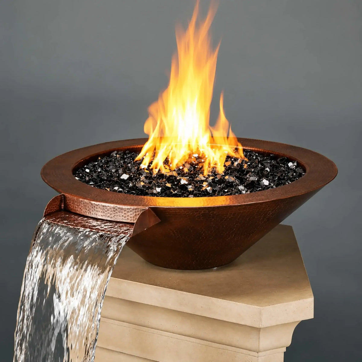 Flame Havens Bella hammered copper fire and water bowl displayed on a beige stone pillar against a gray background. The angled view shows its broad round rim, tapered sides, and reflective black fire glass beneath tall orange flames. A copper scupper at the front left directs a wide sheet of water over the pillar's edge.