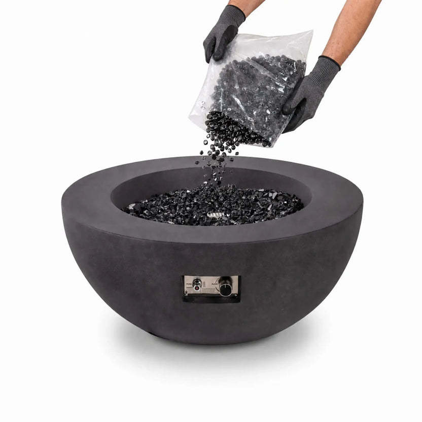 Gloved hands pour Flame Havens reflective 1/2-inch tempered broken black fire glass from a clear bag into a modern round fire pit. The glossy, irregular glass pieces fill the burner area of the dark charcoal bowl, demonstrating safe handling and installation against a clean white background.