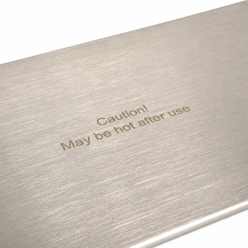Close-up of the Flame Havens Refuge Series 33 x 11-inch stainless steel rectangular burner cover, highlighting its brushed metal grain and engraved safety message reading, “Caution! May be hot after use.” The detailed view shows the cover’s clean finish and clearly visible heat-warning label.