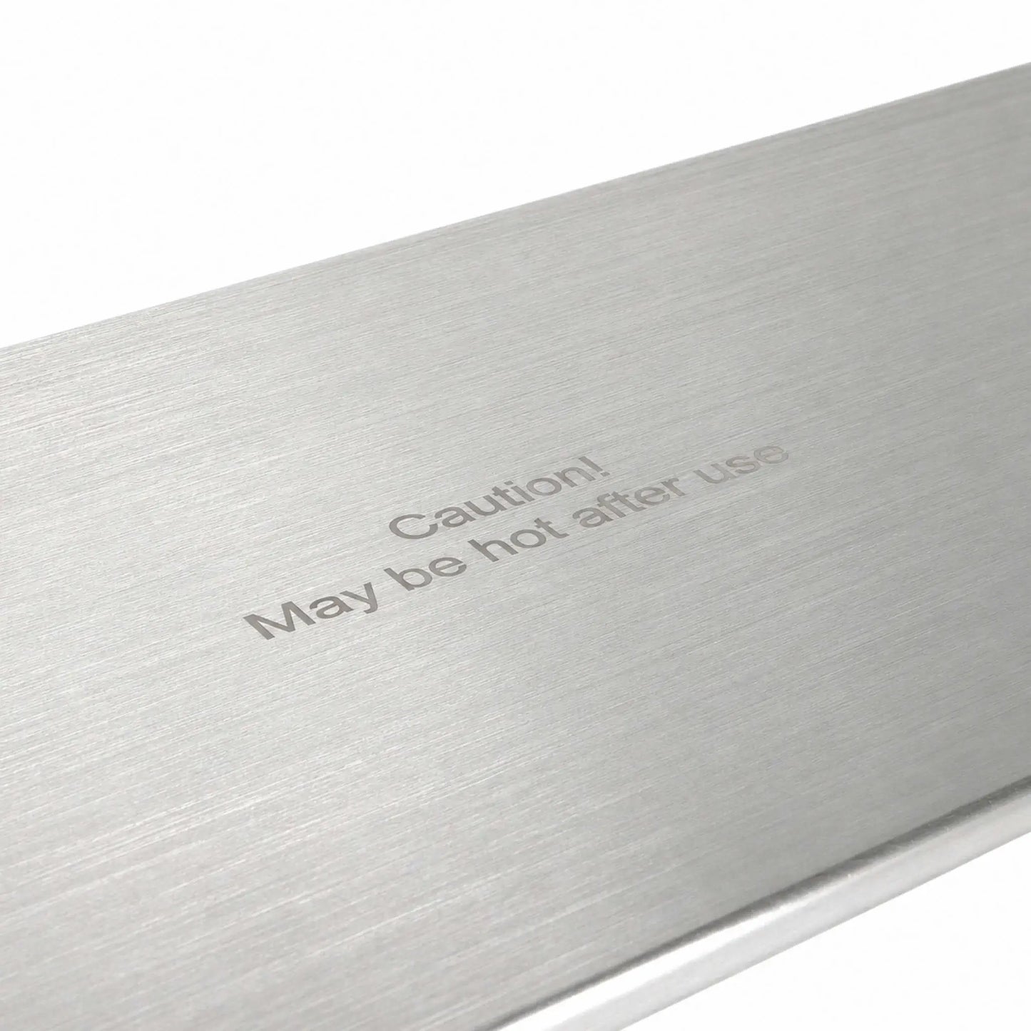 Angled close-up of the Flame Havens Refuge Series 45 x 11-inch stainless steel rectangular burner cover, highlighting its brushed metal surface and centered “Caution! May be hot after use” message. The detailed view shows the fine horizontal grain and clean, finished edge against a white background.