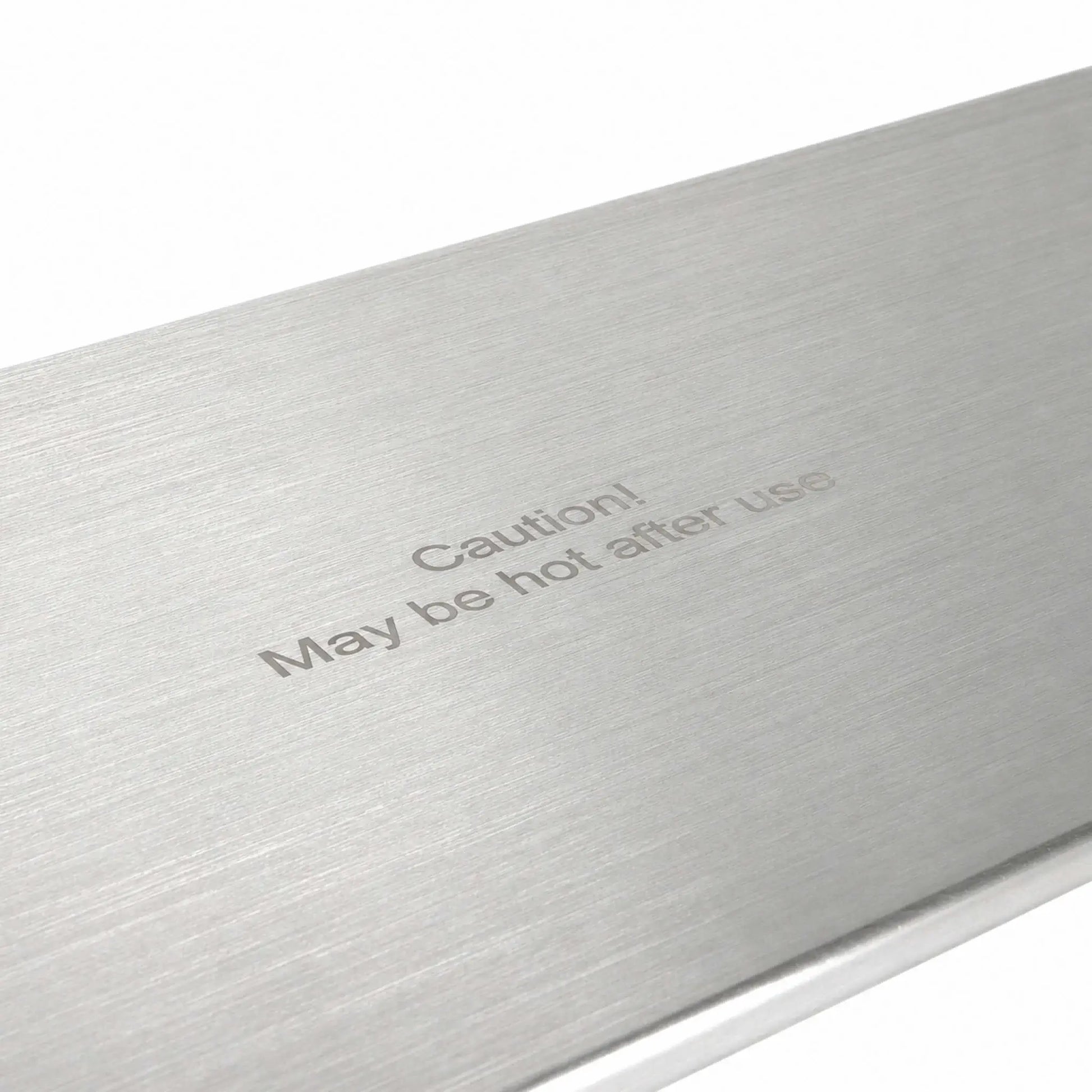 Angled close-up of the Flame Havens Refuge Series 45 x 11-inch stainless steel rectangular burner cover, highlighting its brushed metal surface and centered “Caution! May be hot after use” message. The detailed view shows the fine horizontal grain and clean, finished edge against a white background.