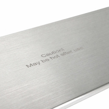 Angled close-up of the Flame Havens Refuge Series 45 x 11-inch stainless steel rectangular burner cover, highlighting its brushed metal surface and centered “Caution! May be hot after use” message. The detailed view shows the fine horizontal grain and clean, finished edge against a white background.