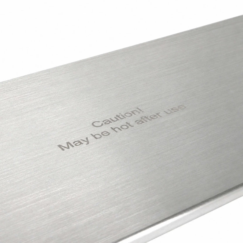 Angled close-up of the Flame Havens Refuge Series 45 x 11-inch stainless steel rectangular burner cover, highlighting its brushed metal surface and centered “Caution! May be hot after use” message. The detailed view shows the fine horizontal grain and clean, finished edge against a white background.