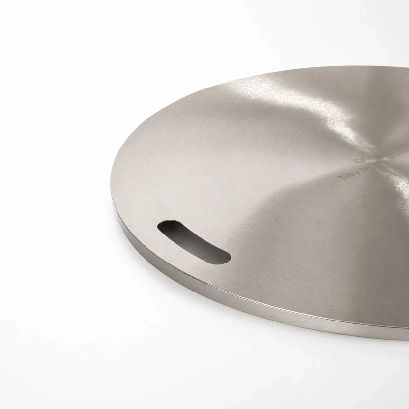 Close-up of the left handle on the Flame Havens Refuge Series 24-inch round stainless steel burner cover. The image highlights the smooth recessed hand slot, brushed metal finish, rounded circular edge, and part of the engraved heat caution near the center.