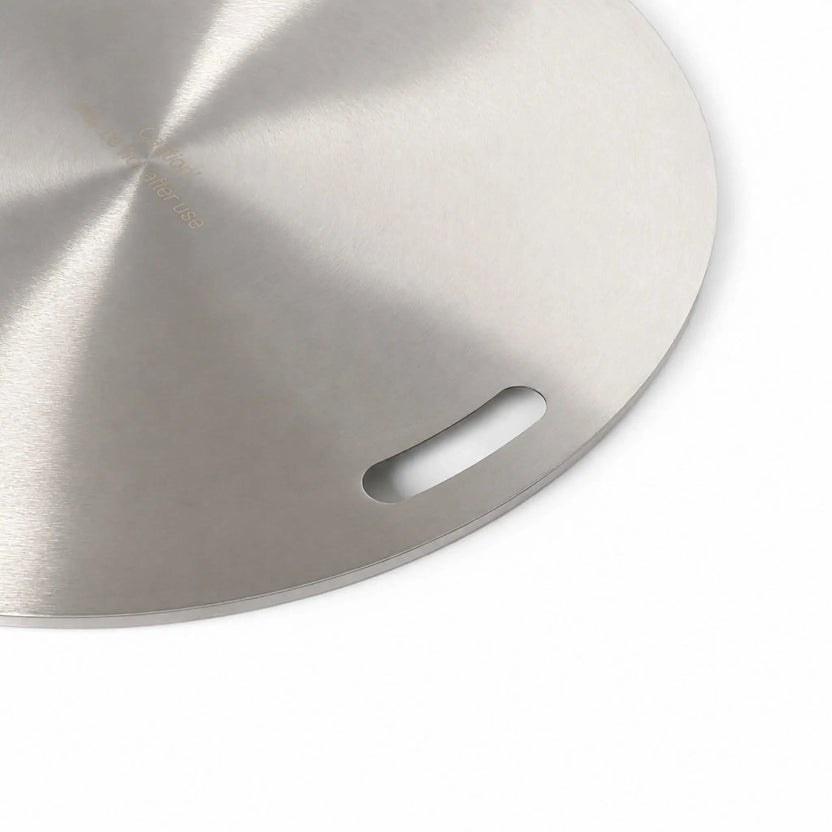 Close-up of the right handle on the Flame Havens Refuge Series 24-inch round stainless steel burner cover. The brushed circular surface, recessed hand slot, finished outer edge, and partial “Caution! May be hot after use” engraving are visible against a white background.