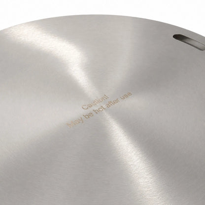 Close-up of the Flame Havens Refuge Series 24-inch round stainless steel burner cover. The brushed metal surface shows its circular grain, one recessed handle opening near the edge, and the engraved safety message, “Caution! May be hot after use.”