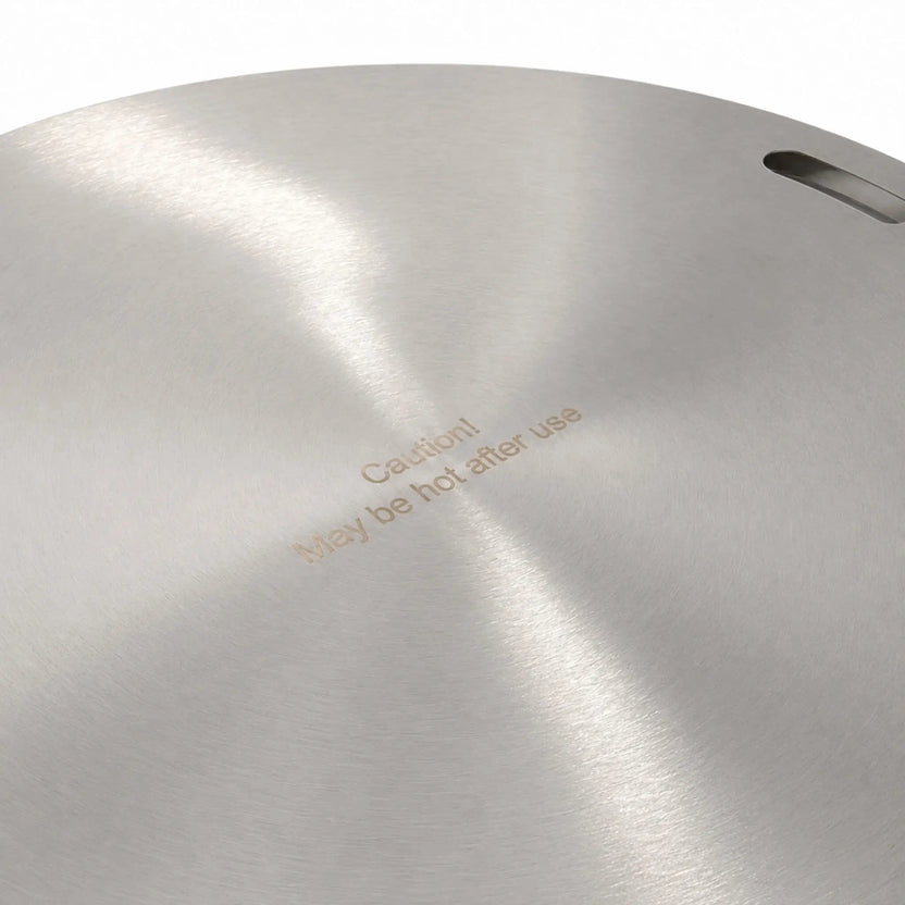 Close-up of the Flame Havens Refuge Series 24-inch round stainless steel burner cover. The brushed metal surface shows its circular grain, one recessed handle opening near the edge, and the engraved safety message, “Caution! May be hot after use.”