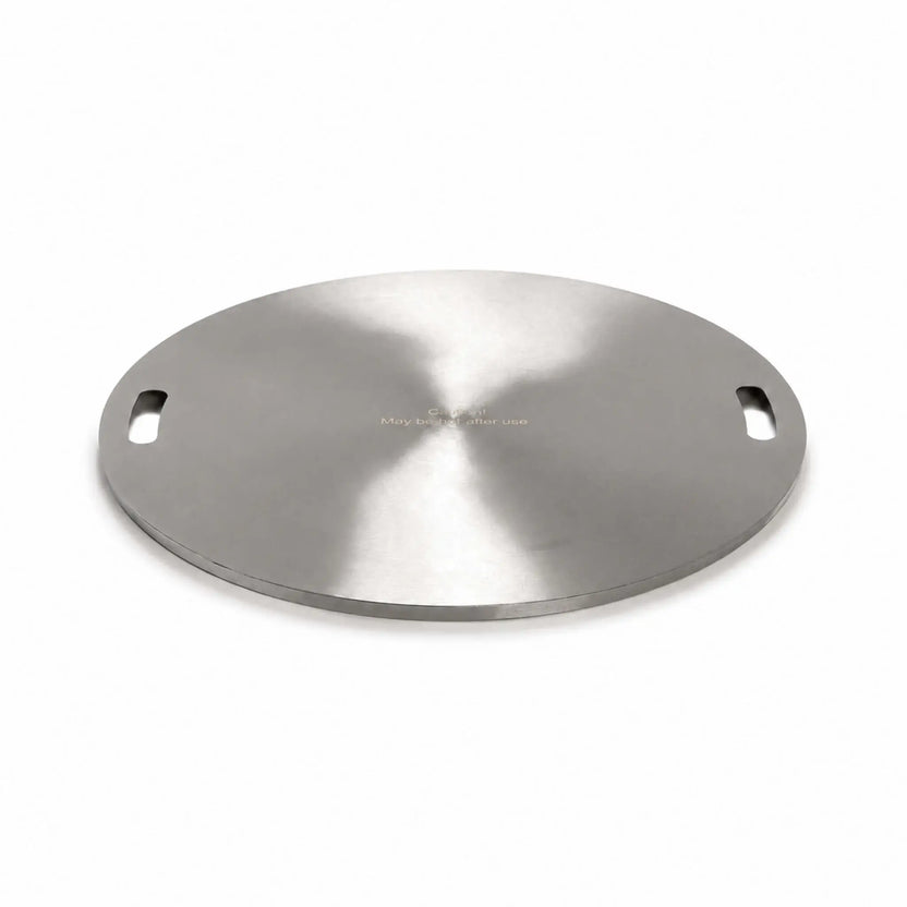 Angled product view of the Flame Havens Refuge Series 24-inch round stainless steel burner cover on a white background. The circular cover has a brushed finish, two recessed carrying handles positioned on opposite sides, a low-profile finished edge, and a centered warning that it may be hot after use.