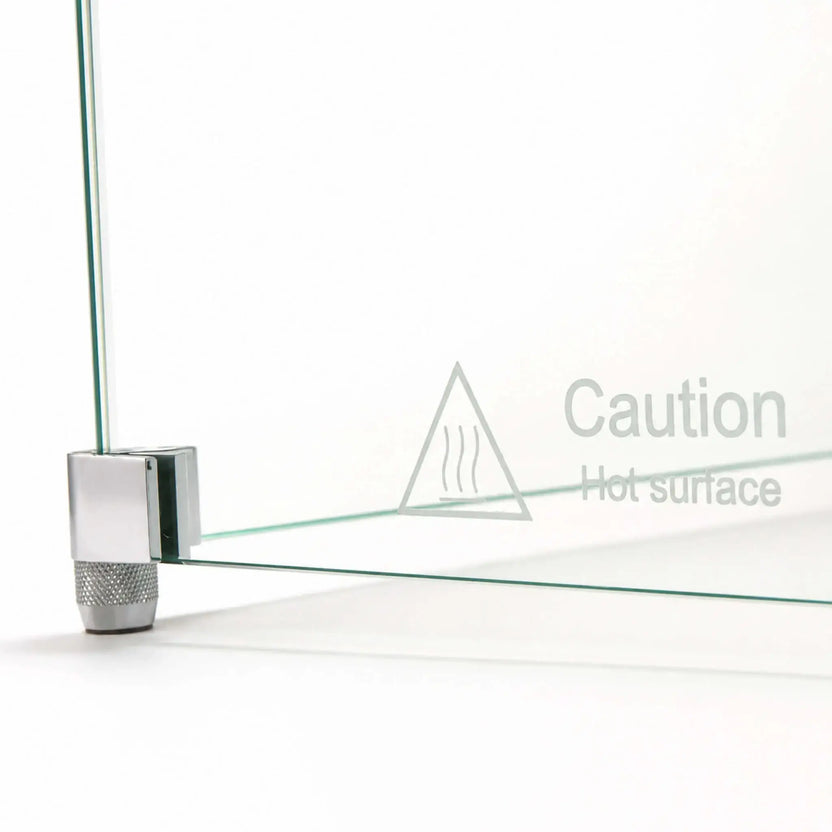 Detailed side view of the Flame Havens Refuge Series 36 x 14 x 7-inch tempered glass wind guard shield corner foot hardware. The image highlights a clear glass panel secured inside a silver metal bracket with a knurled support foot, alongside a triangular heat symbol and “Caution Hot surface” warning.
