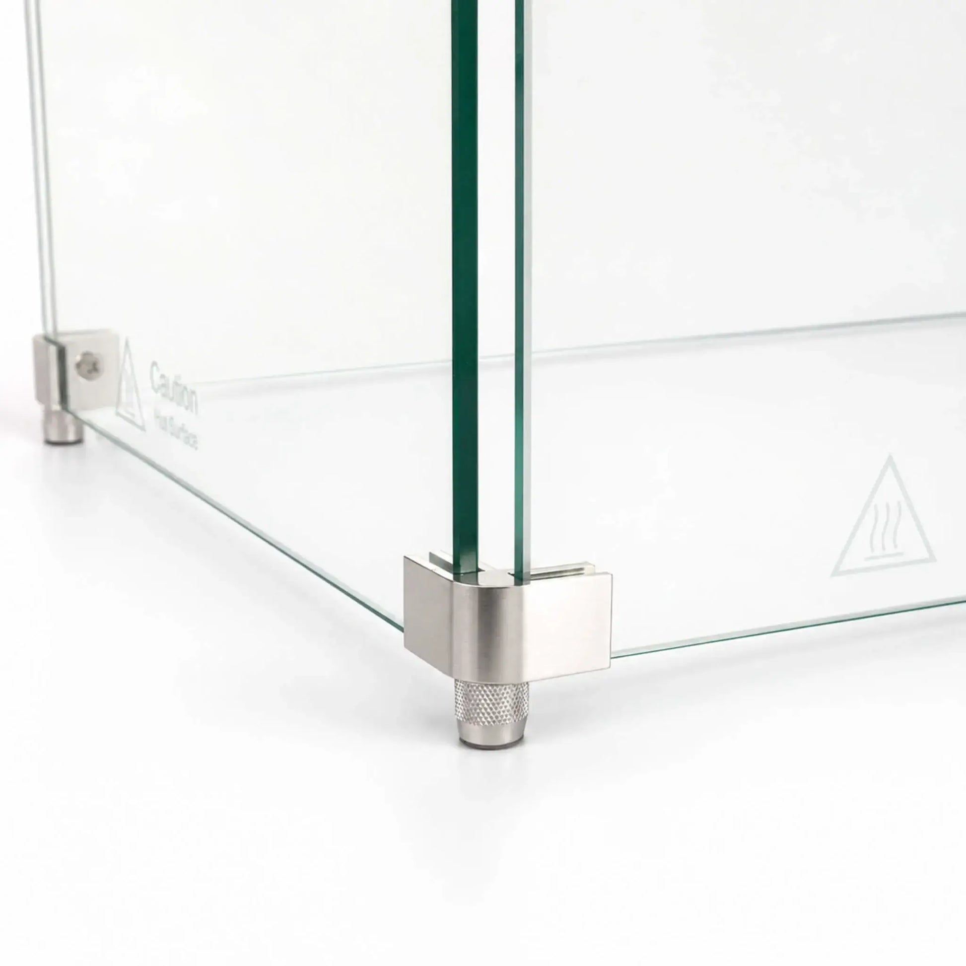Close-up of the corner foot hardware on the Flame Havens Refuge Series 36 x 14 x 7-inch rectangular tempered glass wind guard shield. Two clear glass panels meet inside a brushed stainless steel corner bracket supported by a textured cylindrical foot, with a hot-surface warning visible on the glass.