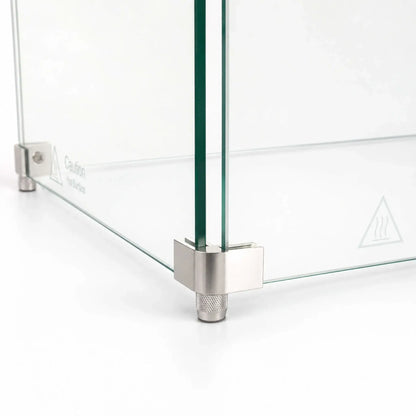 Close-up of the corner foot hardware on the Flame Havens Refuge Series 36 x 14 x 7-inch rectangular tempered glass wind guard shield. Two clear glass panels meet inside a brushed stainless steel corner bracket supported by a textured cylindrical foot, with a hot-surface warning visible on the glass.