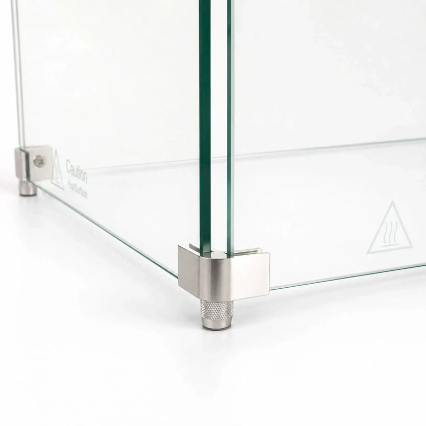 Close-up of the corner foot hardware on the Flame Havens Refuge Series 36 x 14 x 7-inch rectangular tempered glass wind guard shield. Two clear glass panels meet inside a brushed stainless steel corner bracket supported by a textured cylindrical foot, with a hot-surface warning visible on the glass.
