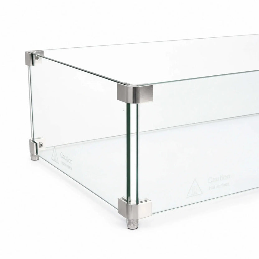 Close-up perspective of the Flame Havens Refuge Series 36 x 14 x 7-inch rectangular tempered glass wind guard shield, showing clear panels joined at the corners with brushed silver metal hardware. The image highlights the upper connectors, lower brackets, adjustable feet, polished edges, and hot-surface safety markings.
