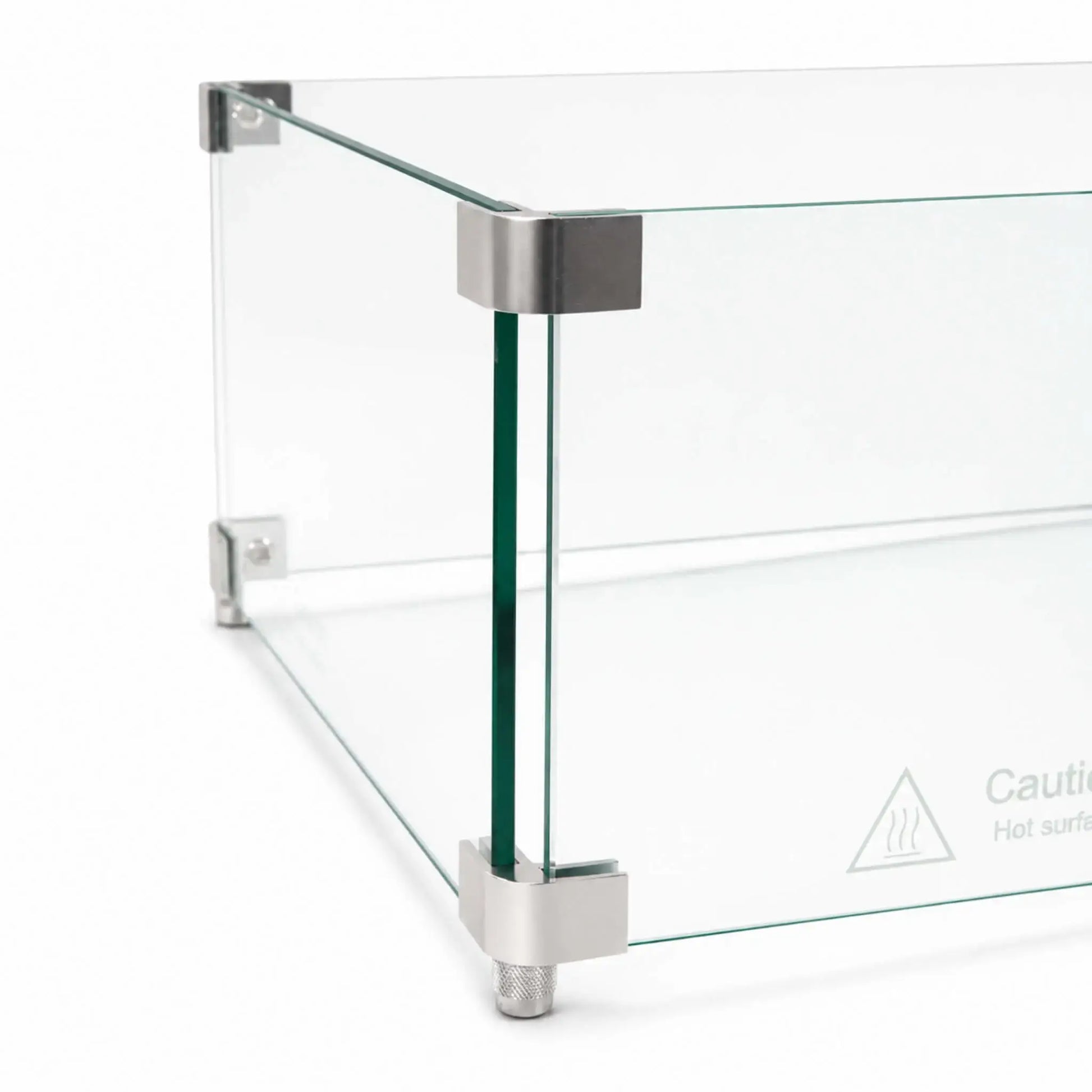 Detailed corner view of the Flame Havens Refuge Series 36 x 14 x 7-inch tempered glass wind guard shield. Two clear glass panels meet inside smooth silver upper and lower corner brackets, with a textured support foot below and a “Caution Hot surface” symbol printed near the bottom of the glass.