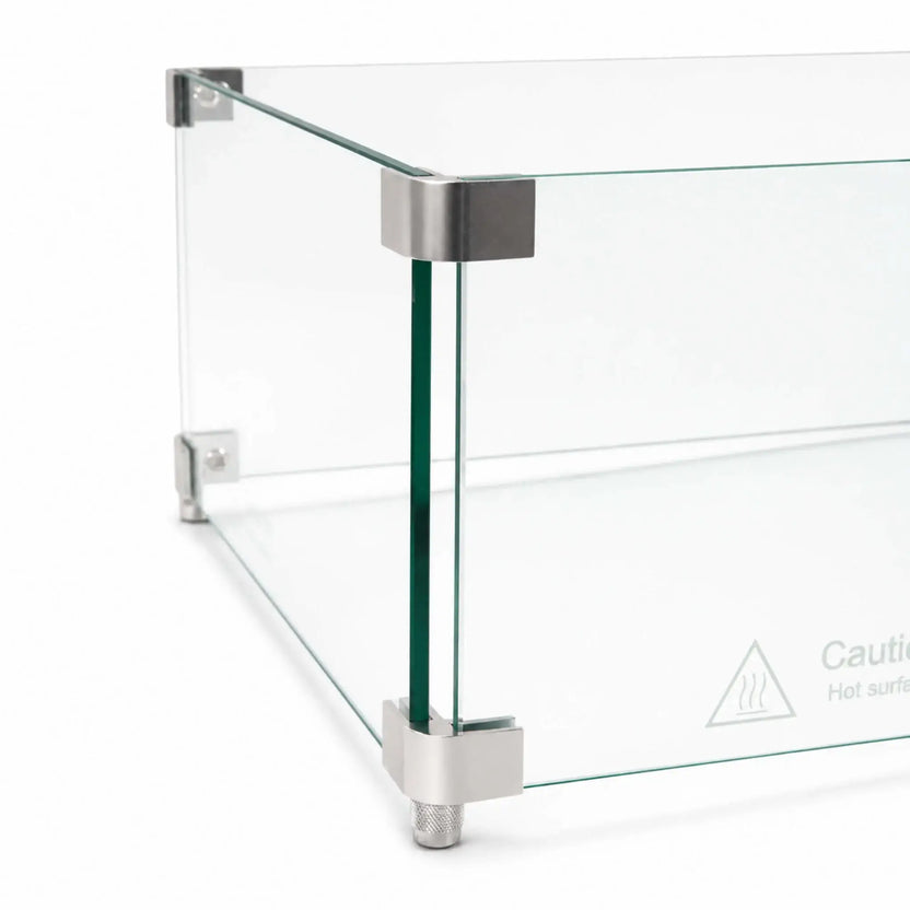 Detailed corner view of the Flame Havens Refuge Series 36 x 14 x 7-inch tempered glass wind guard shield. Two clear glass panels meet inside smooth silver upper and lower corner brackets, with a textured support foot below and a “Caution Hot surface” symbol printed near the bottom of the glass.