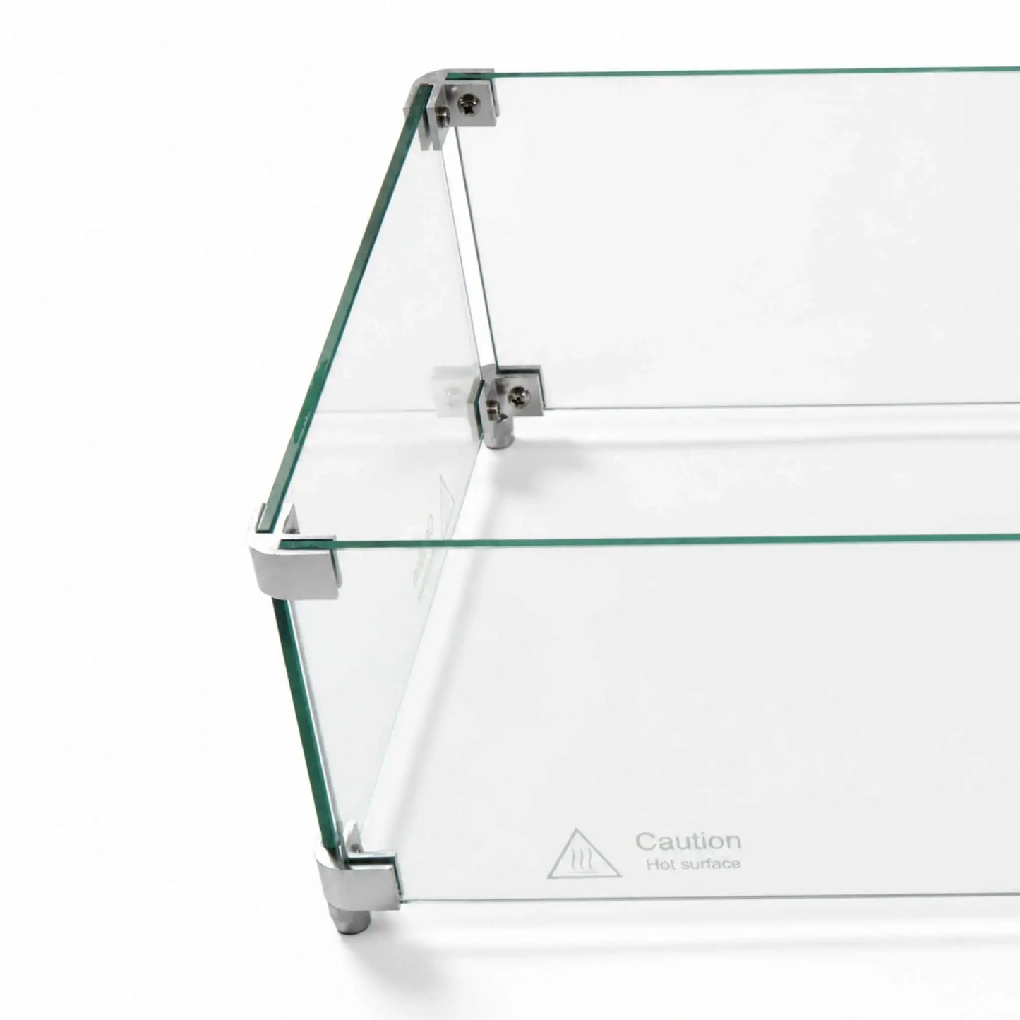 Angled overhead close-up of the Flame Havens Refuge Series 36 x 14 x 7-inch rectangular tempered glass wind guard shield. Clear glass panels are joined by silver corner hardware with visible screws and supporting feet, while a “Caution Hot surface” marking appears near the lower edge.