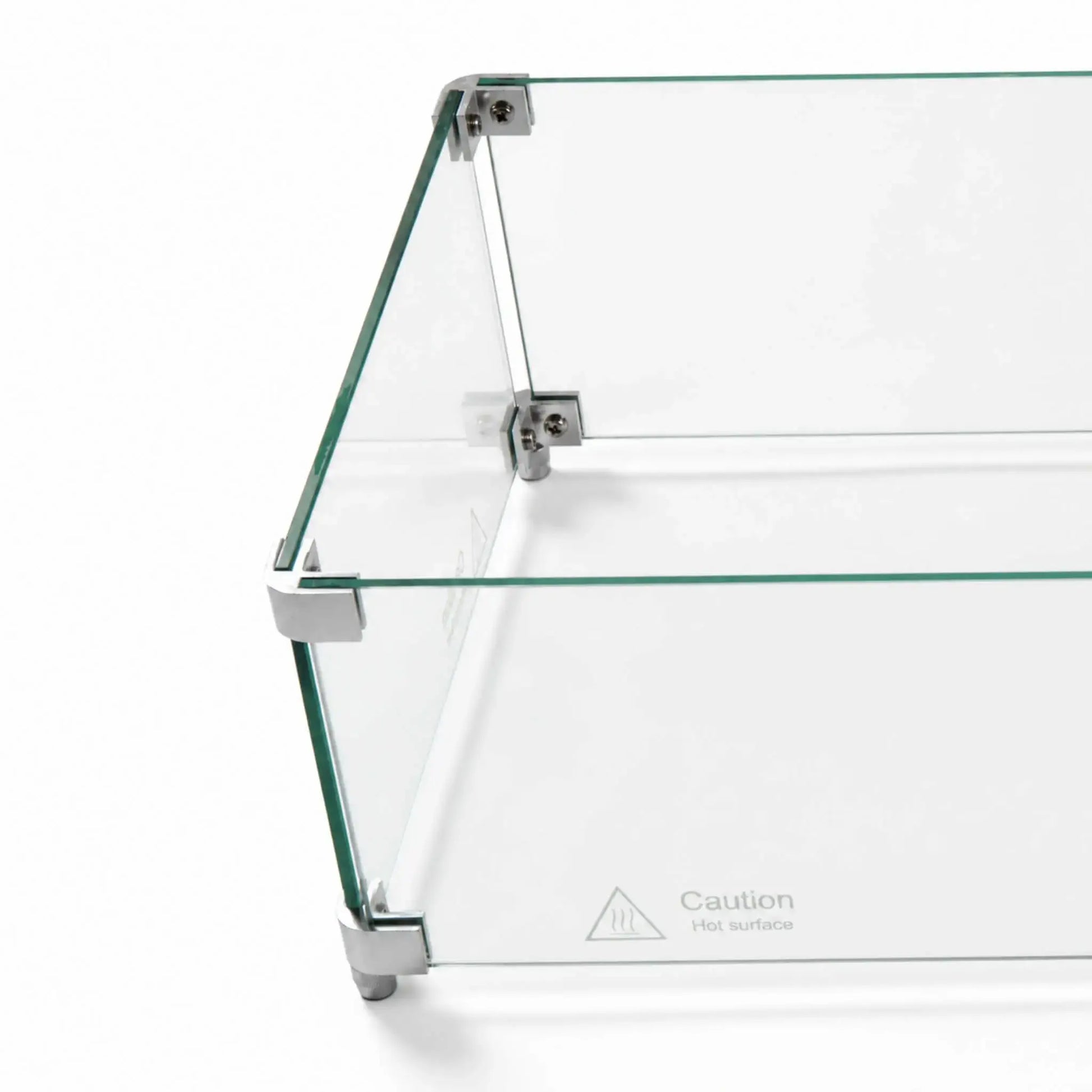 Angled overhead close-up of the Flame Havens Refuge Series 36 x 14 x 7-inch rectangular tempered glass wind guard shield. Clear glass panels are joined by silver corner hardware with visible screws and supporting feet, while a “Caution Hot surface” marking appears near the lower edge.
