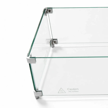 Angled overhead close-up of the Flame Havens Refuge Series 36 x 14 x 7-inch rectangular tempered glass wind guard shield. Clear glass panels are joined by silver corner hardware with visible screws and supporting feet, while a “Caution Hot surface” marking appears near the lower edge.