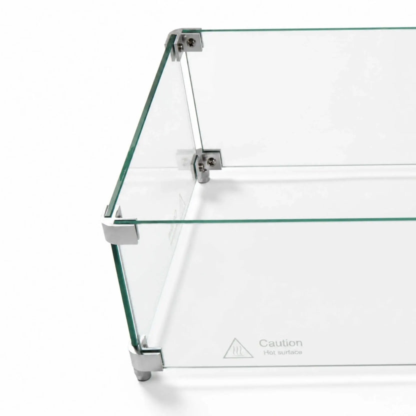 Angled overhead close-up of the Flame Havens Refuge Series 36 x 14 x 7-inch rectangular tempered glass wind guard shield. Clear glass panels are joined by silver corner hardware with visible screws and supporting feet, while a “Caution Hot surface” marking appears near the lower edge.