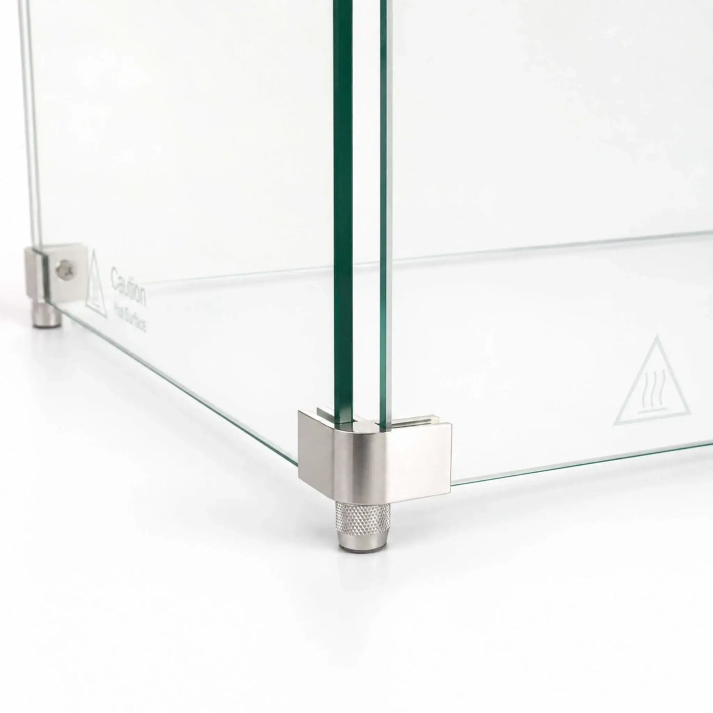Close-up of the feet hardware on the Flame Havens Refuge Series 48 x 14 x 7-inch rectangular tempered glass wind guard. A brushed stainless steel corner bracket holds two clear glass panels above a textured cylindrical foot, with the glass edges and hot-surface caution symbol visible.