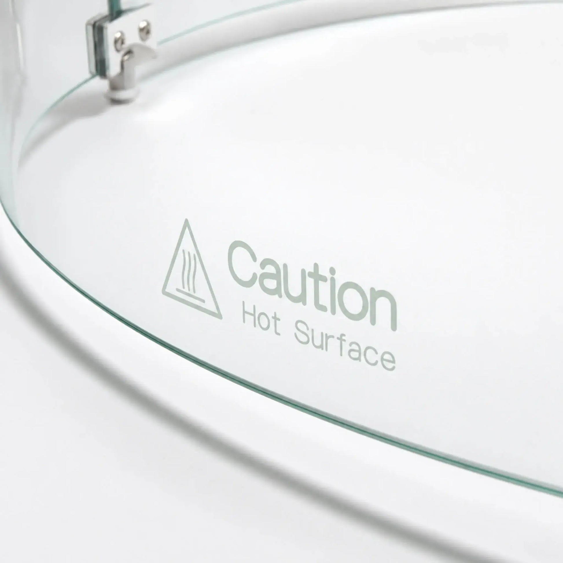 Close-up of the “Caution Hot Surface” warning printed on the Flame Havens Refuge Series round tempered glass wind guard shield. The 28-inch-diameter by 7-inch-tall accessory shows its curved clear glass edge, heat warning symbol, and part of the silver connecting hardware.
