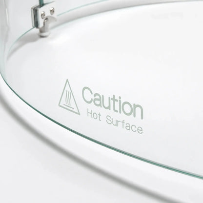Close-up of the “Caution Hot Surface” warning printed on the Flame Havens Refuge Series round tempered glass wind guard shield. The 28-inch-diameter by 7-inch-tall accessory shows its curved clear glass edge, heat warning symbol, and part of the silver connecting hardware.